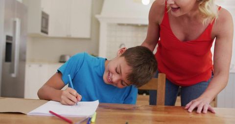 Smiling boy drawing at kitchen table while mother watching and encouraging creativity