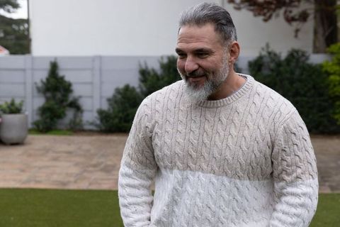 Mature man wearing beige cable-knit sweater smiling standing on lawn in backyard garden