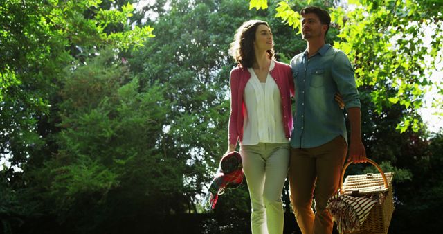 A couple carrying a picnic basket is walking through a green forest. They are enjoying a sunny day, surrounded by lush foliage. This image can be used for themes related to nature, outdoor activities, leisure, romance, and lifestyle advertisements. Ideal for promoting travel destinations, picnic spots, relationship tips, and relaxation products.