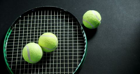 Close-up of bright green tennis balls placed on a stringed racket against a dark background, emphasizing the texture and arrangement. Suitable for themes related to sports, fitness, or tennis activities. Great for use in advertisements for tennis tutorials, equipment retailers, or sports-themed articles.