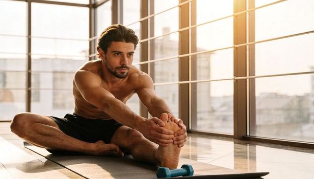Muscular man stretching hamstring on yoga mat in sunlit urban studio with dumbbell