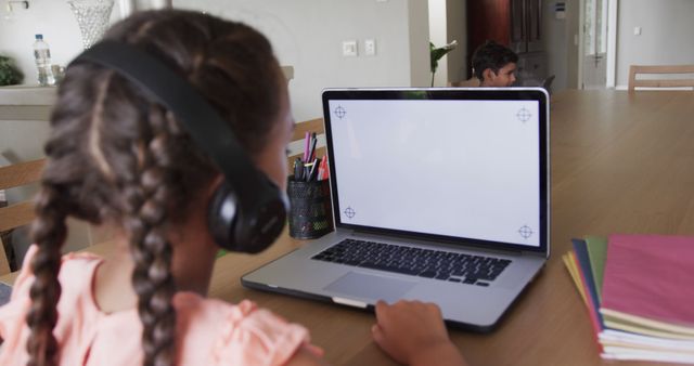 A young girl with braided hair is using headphones while studying on her laptop at a wooden table at home. A boy is in the background. Ideal for themes related to online learning, distance education, homeschooling, or children studying.
