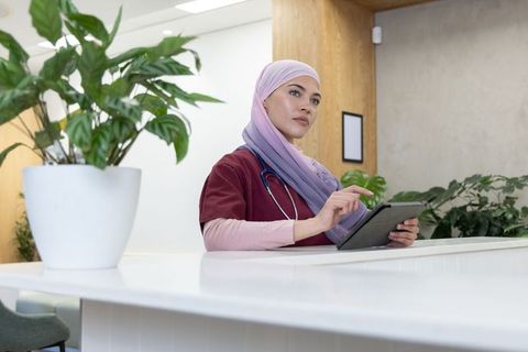 Female nurse wearing hijab using tablet at clinic reception with green plants