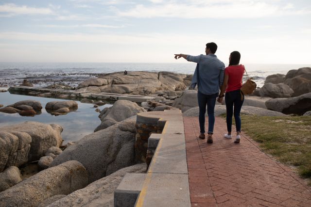 Biracial couple enjoying a walk by the rocky seashore on a sunny day, holding hands and bonding. Ideal for use in travel blogs, relationship articles, and lifestyle magazines. Perfect for promoting outdoor activities, romantic getaways, and nature exploration.