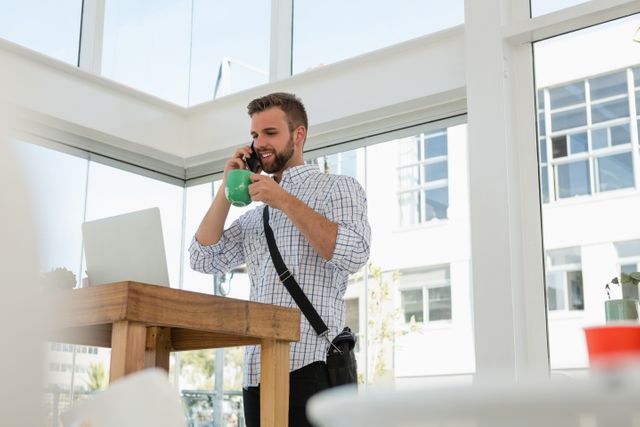 Businessman in casual attire talking on mobile phone while holding a coffee mug at a standing desk in a modern office. Ideal for use in articles or advertisements about modern work environments, productivity, remote work, or office technology.