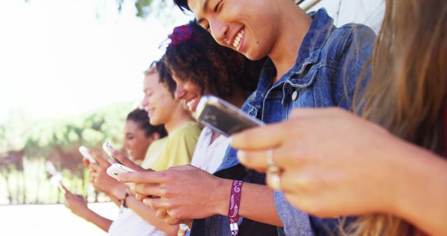 A group of smiling teens standing outdoors, all using their smartphones. This image is perfect for themes related to technology use among young people, social media interactions, and youth culture. Suitable for advertisements, blog posts, and articles about modern communication, teen lifestyles, and friendship.