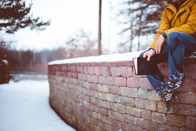Person sitting on a snowy brick wall in winter, holding a book. Wearing a yellow jacket and boots. Ideal for illustrating peaceful winter scenes, outdoor activities in cold weather, solitude, and moments of relaxation during the winter season.
