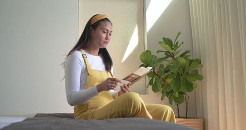 Mid-adult asian woman reading tablet on bed in minimalist bedroom with plant