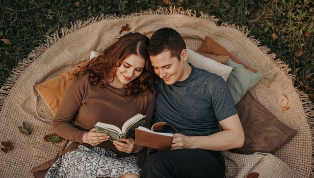 Cozy autumn couple reading on round picnic blanket with cushions and novels in park