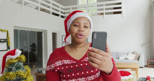 Happy woman wearing Santa hat making video call during cheerful Christmas celebration in a bright, spacious living room. This image can be used for promoting holiday-themed products, advertisements for video call technology, and festive greeting cards showcasing virtual celebrations.