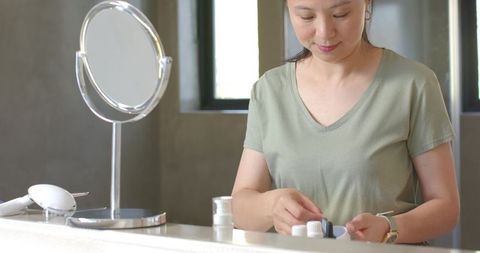 Asian woman arranging skincare jars on bathroom vanity with mirror and natural light