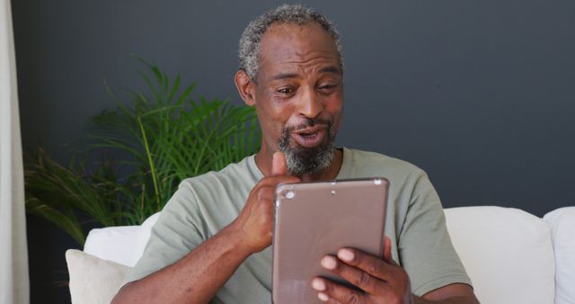 Senior African American man engaging in a video call on a tablet while relaxing on a sofa in a home environment. Perfect for use in advertisements or articles related to technology adoption by older adults, communication tools, staying connected with family or friends, and promoting video call applications.