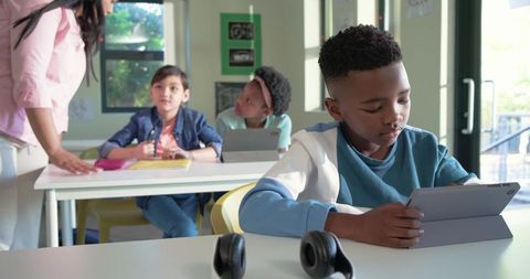 African american boy using tablet while classmates collaborating with teacher in classroom
