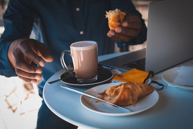 African American man sitting at a table with a laptop, enjoying a cup of coffee and a croissant. Ideal for illustrating concepts of remote work, digital nomad lifestyle, business, casual dining, and urban living. Suitable for use in articles, blogs, advertisements, and social media posts related to work-life balance, productivity, and modern work environments.