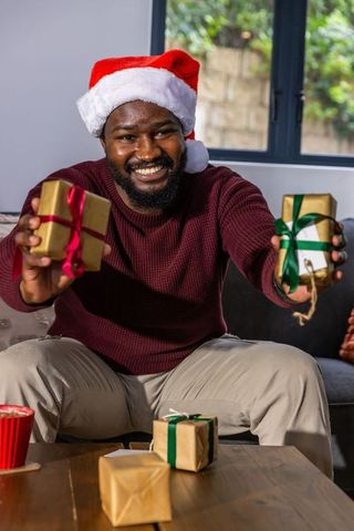 African american man wearing santa hat presenting wrapped gifts and smiling on cozy couch