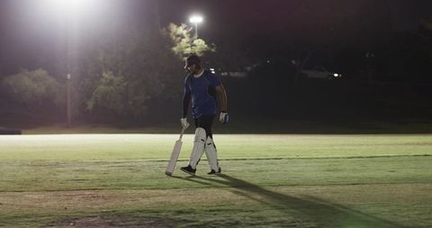 Cricket batter walking across illuminated pitch at night holding bat wearing pads, helmet