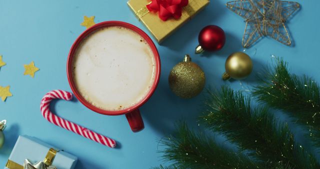 Warm cup of hot chocolate surrounded by Christmas decorations including wrapped gifts, candy cane, ornaments, and greenery on a blue background. Perfect for holiday promotions, seasonal greetings, and festive content.