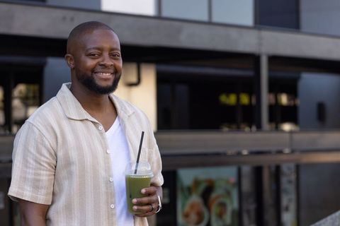 African american man smiling on balcony holding green smoothie summer lifestyle portrait