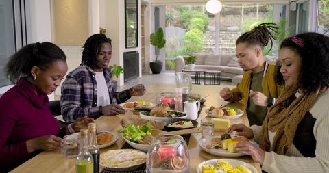 Multicultural friends enjoying home-cooked meal at wooden dining table in open-plan home