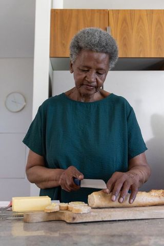 Senior african woman slicing baguette in home kitchen preparing buttered bread