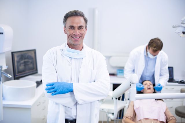 Dentist wearing gloves and white coat standing confidently with arms crossed while patient receives treatment in background. Useful for healthcare campaigns, dental care promotions, and dental clinic marketing materials.