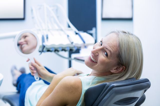 Female patient smiling while holding a mirror in a dental clinic. She is sitting in a dental chair, looking pleased with the dental care she has received. This image can be used for promoting dental services, oral health awareness, dental hygiene products, and healthcare advertisements.