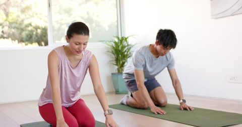 Diverse couple practicing yoga kneeling on green mats by window in bright home studio