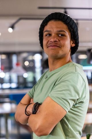 Young man standing arms crossed wearing beanie and green tee in coworking cafe