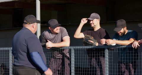 Sunlit baseball teammates leaning on dugout railing chatting, holding glove, wearing caps