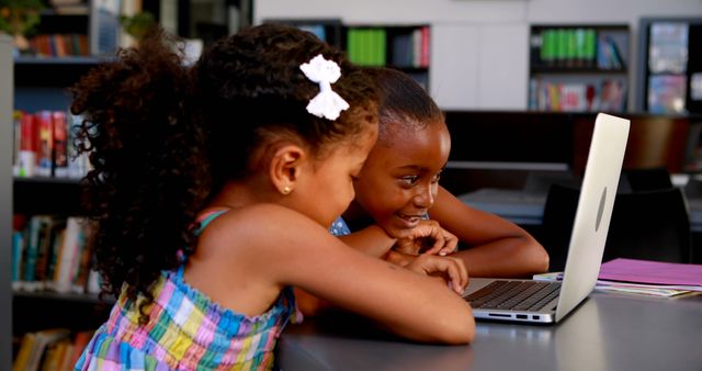 Two young girls smiling and working on a laptop in a library. Image ideal for illustrating concepts of early education, friendship in learning, and the integration of technology in traditional learning environments.