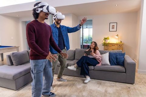Diverse friends playing virtual reality headsets in modern living room, woman using tablet