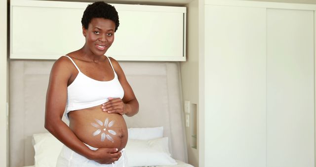 Smiling african american pregnant woman with flower on her belly at home, copy space. Pregnancy, motherhood, domestic life and wellbeing concept, unaltered.