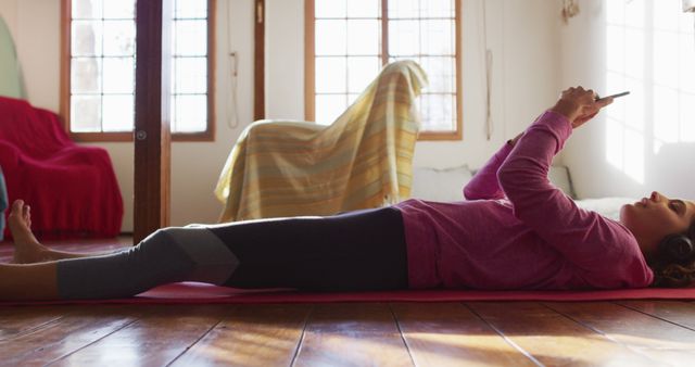 Young woman lying on yoga mat using smartphone in cozy, sunlit room. Could be useful for promoting self-care, at-home fitness, or relaxation tips. Highlights casual, everyday lifestyle and technology usage.
