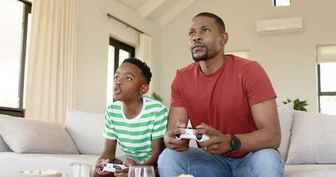 African American father and son playing video games on sofa bonding in bright living room