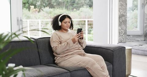 Mid-adult African American woman lounging on dark sofa wearing headphones and using smartphone