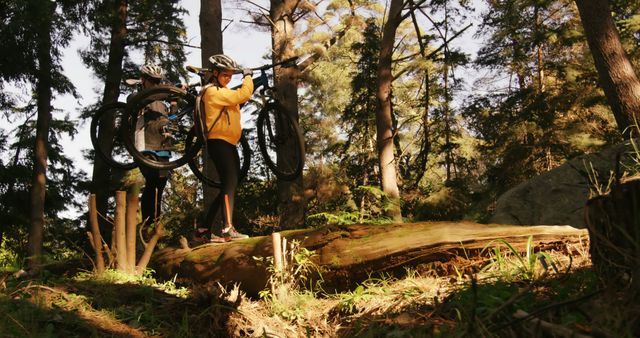 A young Caucasian woman in athletic gear is carrying her mountain bike over a fallen tree on a forest trail, with copy space. Her determination and the challenging outdoor environment emphasize the adventurous spirit of mountain biking.