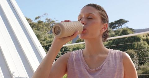 Woman drinking from beige reusable bottle on sunlit balcony overlooking green hillside