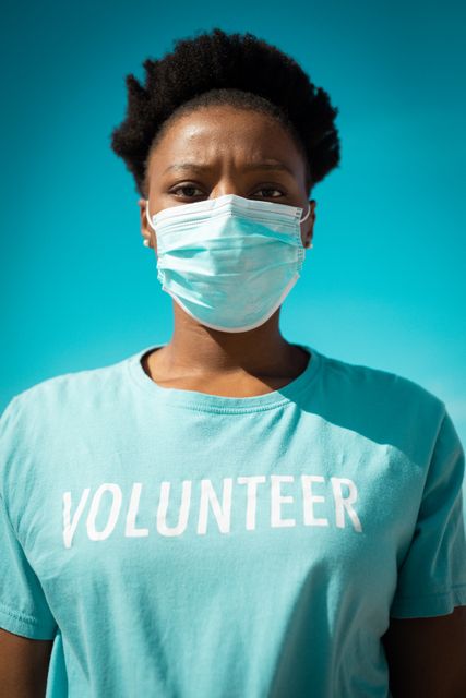 African american woman wearing volunteer t shirts and face mask, looking at camera. eco conservation volunteers, beach clean-up during coronavirus covid 19 pandemic.