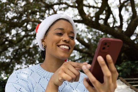 African american woman wearing santa hat smiling while tapping smartphone beneath tree