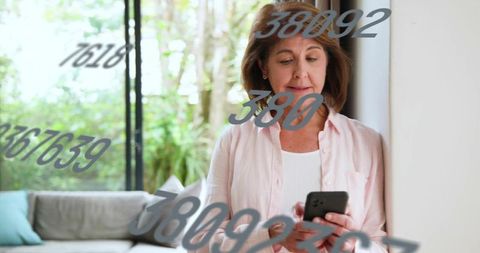 Senior woman holding smartphone by window with floating numbers overlay in bright home