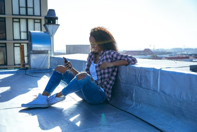 Side view of a hip young biracial woman wearing checked shirt, jeans and white trainers, using her smartphone, smiling and sitting on an urban rooftop with buildings in the background.