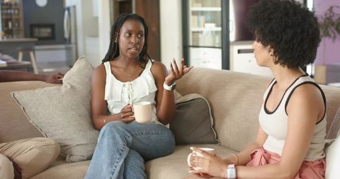 African American women chatting on sofa holding mugs, enjoying cozy home conversation