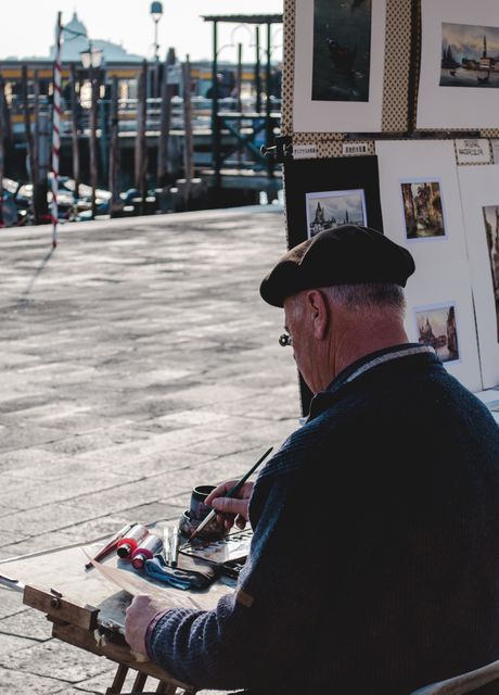 Senior artist painting outdoor by waterfront, captured from behind. Featuring a man working on his artwork using brushes and paints. Paintings and sketches displayed on a stand next to him, highlighting a vibrant and scenic setting near a pier. Suitable for concepts related to art, creativity, leisure, and street performance. Ideal for use in articles, blogs, or promotional materials focusing on art, elderly hobbies, or outdoor activities in a picturesque location.