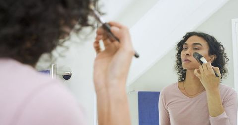 Nonbinary person applying foundation with brush at bathroom mirror, grooming and beauty