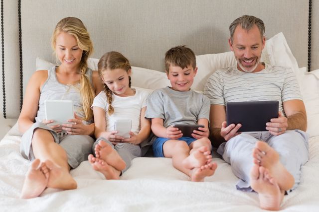 Smiling parents and kids using mobile phone and digital tablet on bed in bedroom