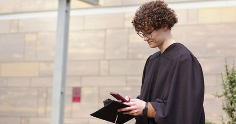 College graduate checking phone while holding mortarboard outside campus courtyard celebration