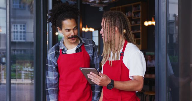 Two male restaurant workers wearing red aprons are discussing orders on a digital tablet. They are standing near the entrance of a cafe and appear to be collaborating. The background shows a well-lit interior with tables, chairs, and shelves. This image can be used for themes related to teamwork, hospitality industry, technology in the workplace, effective employee communication, and modern workplaces.