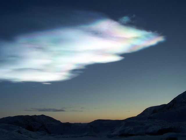 Image shows breathtaking polar stratospheric cloud illuminated at dusk over a remote Arctic landscape. The vibrant colors in the cloud contrast with the darkening sky and mountainous terrains. Ideal for topics related to natural phenomena, atmospheric studies, and remote geography. Suitable for use in educational content, travel magazines, and as background visuals in nature documentaries.