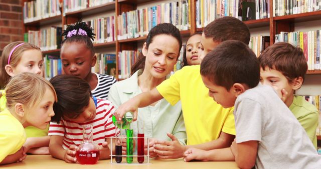 The teacher is sitting with a group of young, diverse children in a library, focusing on a science experiment with test tubes and colorful liquids. This image is suitable for use in educational materials, school brochures, advertisements promoting science programs, or any media highlighting the importance of early childhood education and diverse learning environments.