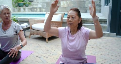 Mature asian and caucasian women practicing gentle yoga and stretching on patio by pool